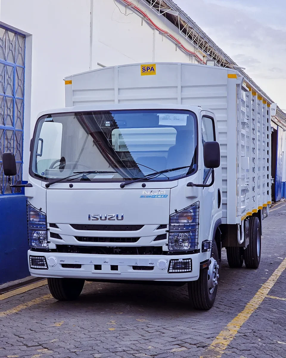 A front-angle view of a white Isuzu NQR Blue Power light-duty truck. It features a high-clearance cargo body built by SPA Industries Ltd, protective metal grilles over the headlights and fog lights, and a clean, industrial white finish. The truck is parked on a paved surface between two white buildings.