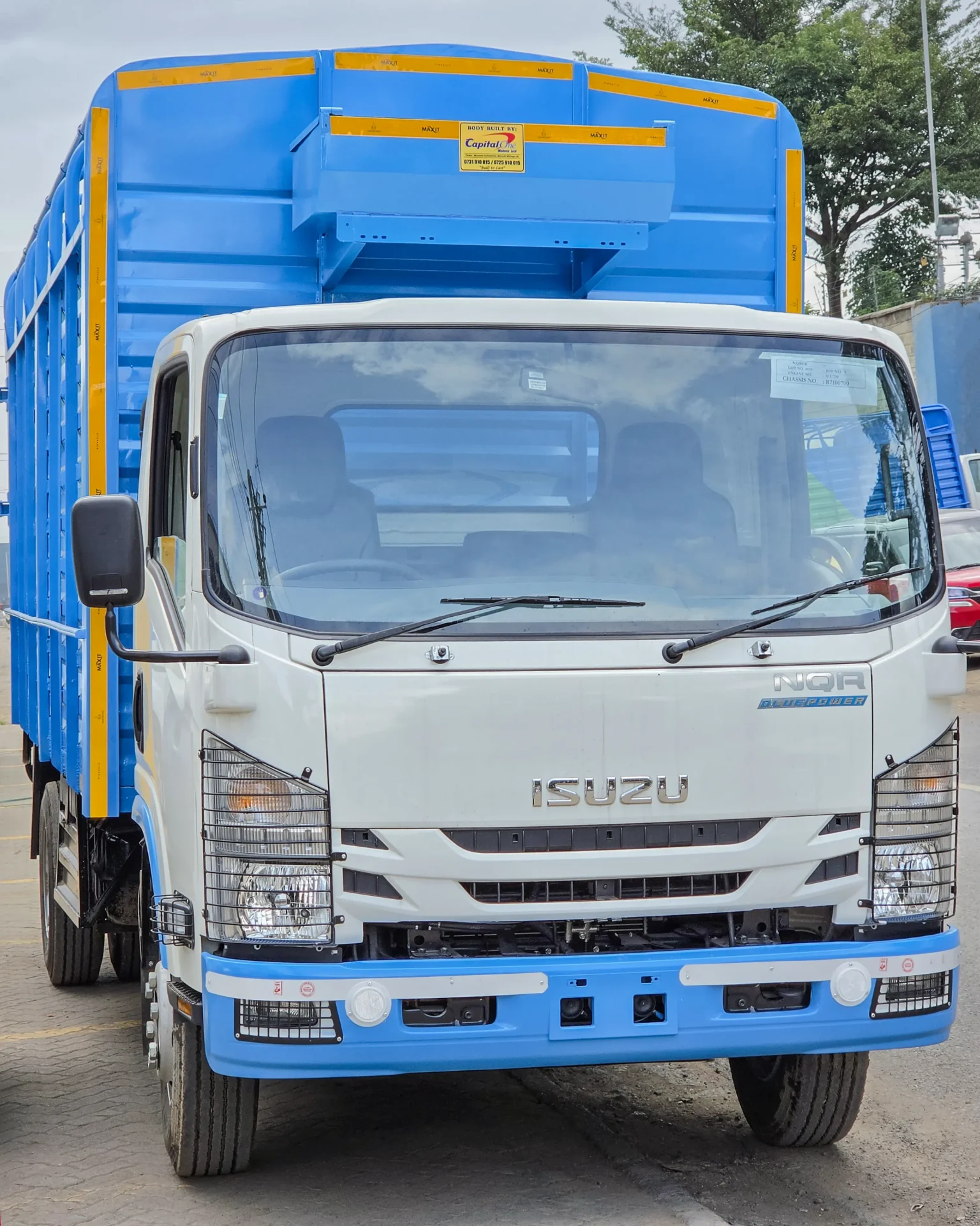 Front view of a white Isuzu NQR truck with a bright blue cargo body and yellow reflective strips, parked outdoors on a paved lot under a cloudy sky.
