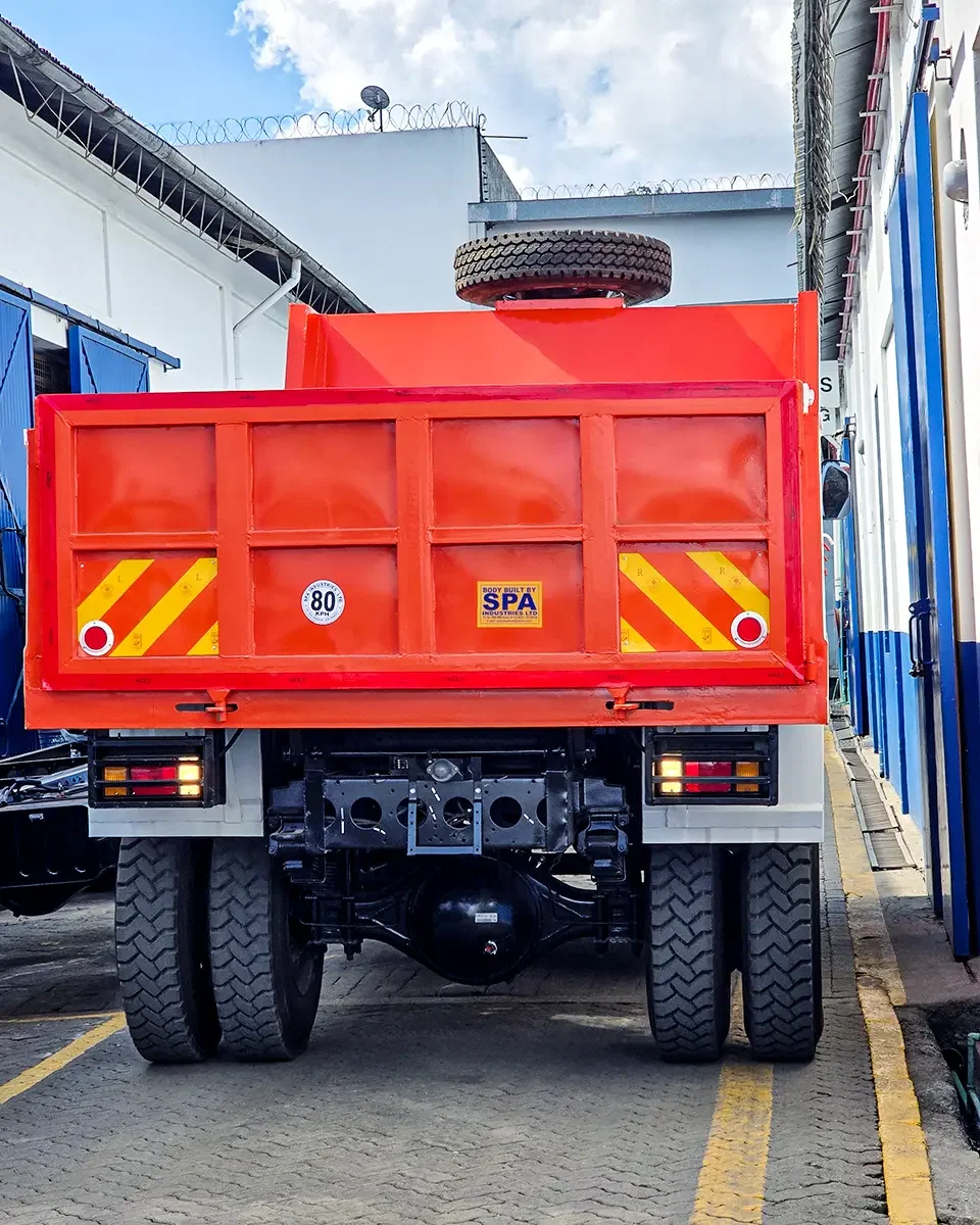 Rear view of an orange Isuzu tipper truck parked in an industrial yard, showing reflective hazard markings, dual rear wheels, and a spare tire mounted on top of the raised dump bed.