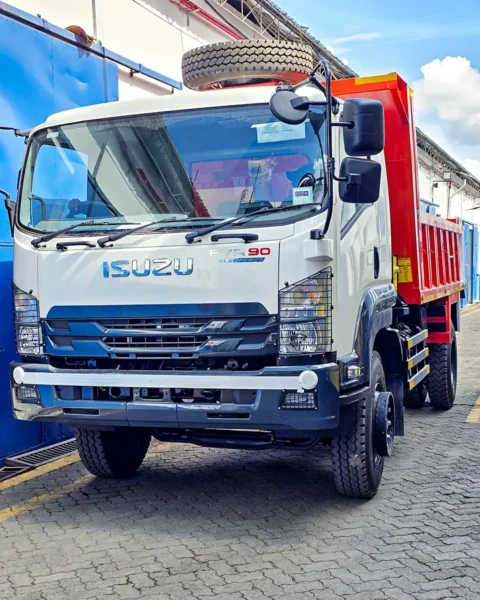 Front-side view of a white Isuzu FVR90 tipper truck with an orange dump body, parked in a paved industrial area, featuring a large mounted spare tire above the cab and heavy-duty front headlights.
