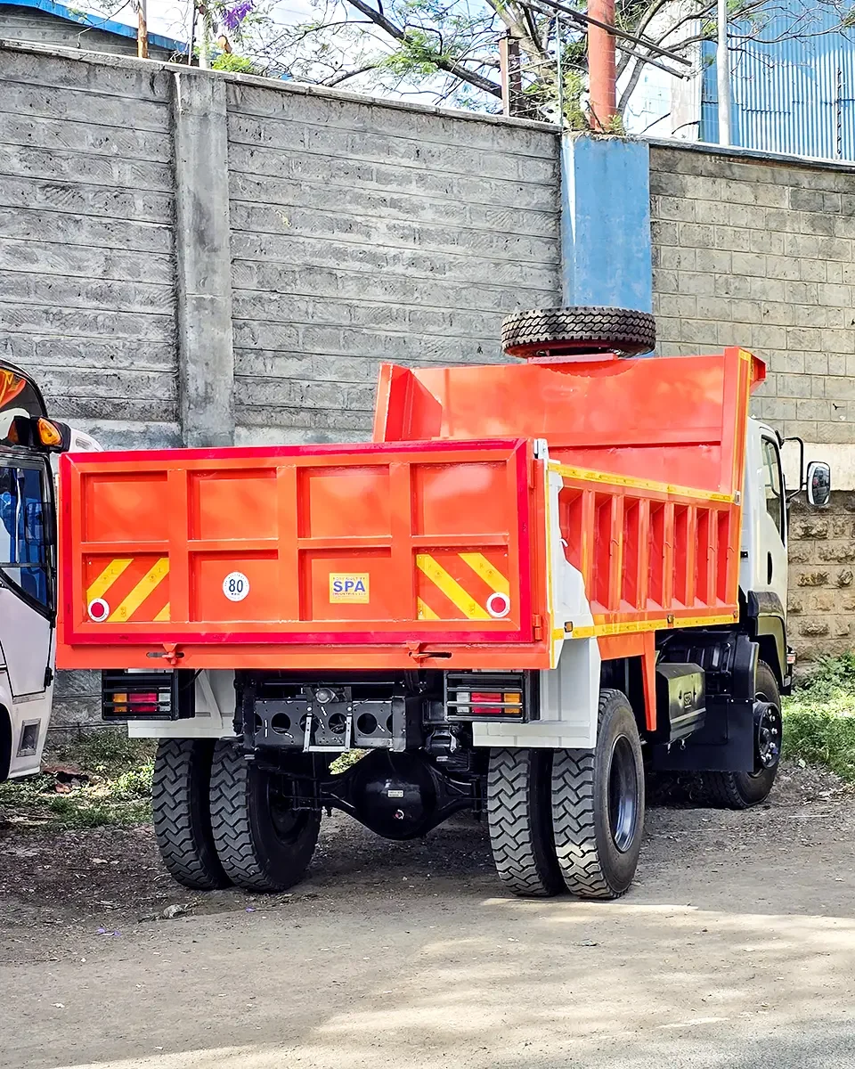 Rear view of an orange dump-body Isuzu tipper truck parked outdoors near a concrete wall, with its dump bed raised slightly and a spare tire mounted above the cab.