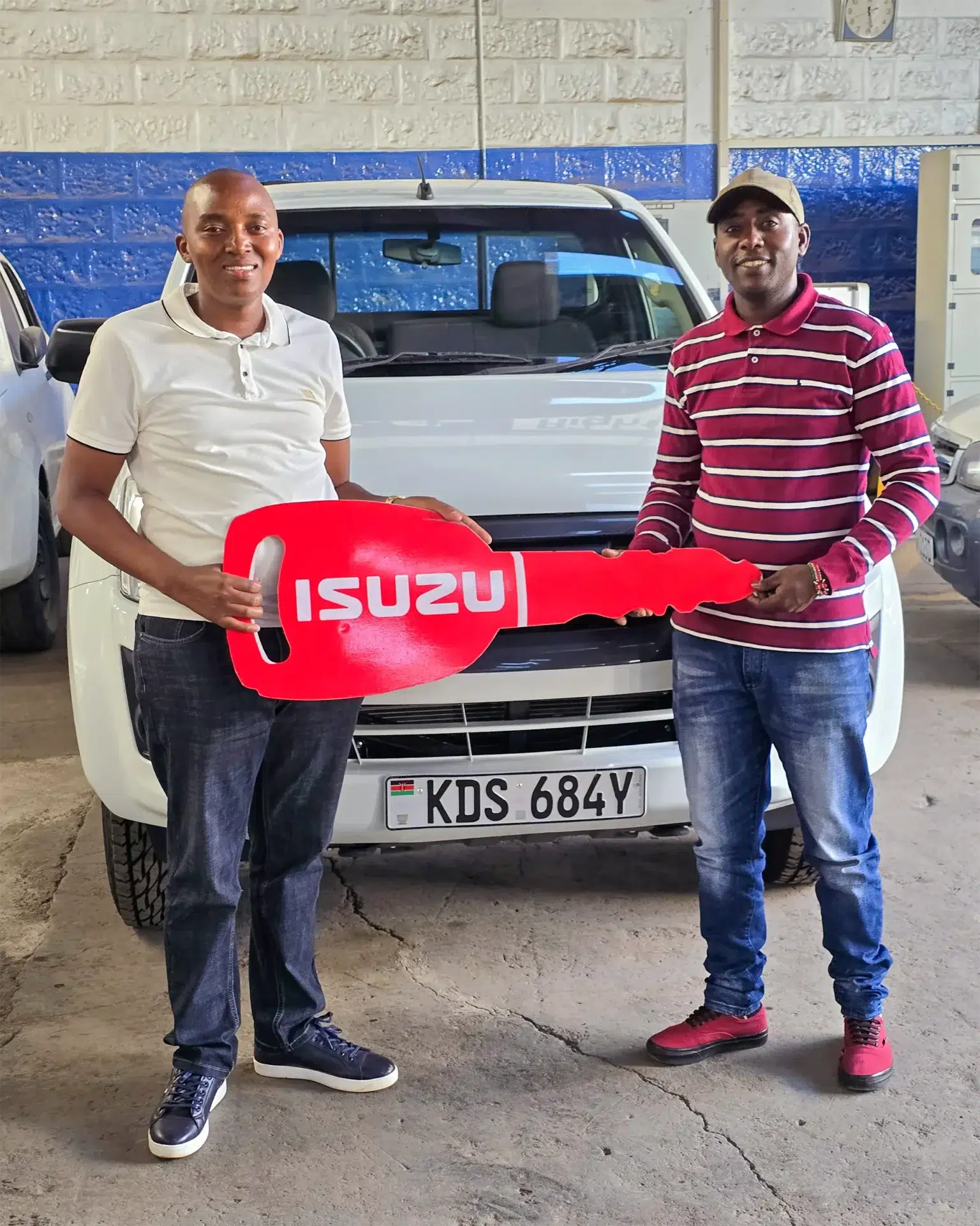 Wang'ombe, a top Isuzu sales representative, handing over a large red Isuzu-branded key to Mr. Kimathi in front of a newly purchased white Isuzu D-Max Single Cab Pick-up with the license plate KDS 684Y, inside a dealership garage.