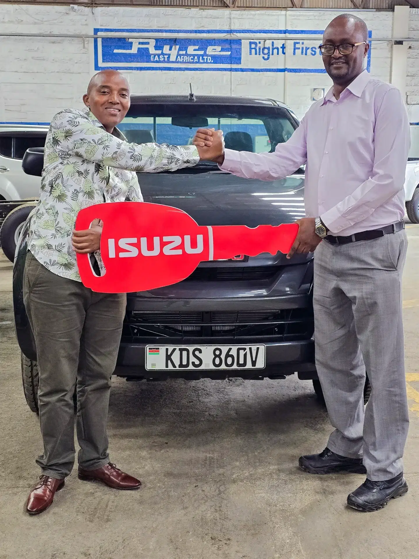 Wang'ombe, a top Isuzu sales representative, handing over a large red Isuzu-branded key to a WELL HOOD EA LTD representative in front of a newly purchased black Isuzu D-Max Single Cab Pick-up with the license plate KDS 86GV, inside a dealership garage.