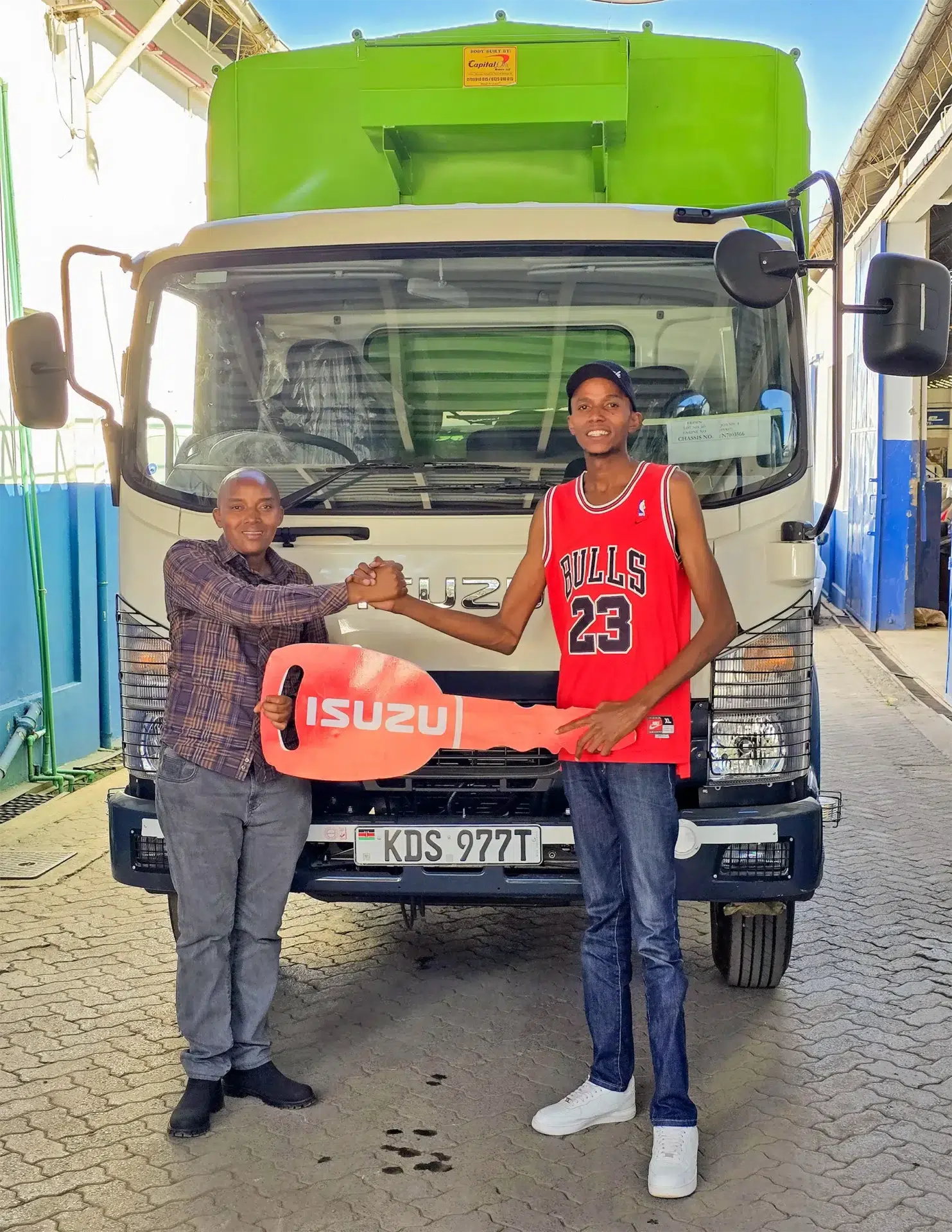 Wang'ombe, a top Isuzu sales representative, handing over a symbolic Isuzu key to Mr. Gideon Wachira in front of a newly purchased Isuzu FRR 90 BLUEPOWER Truck (KDS 977T) with a green cargo box. The two shake hands, celebrating another successful Isuzu truck sale in Kenya.