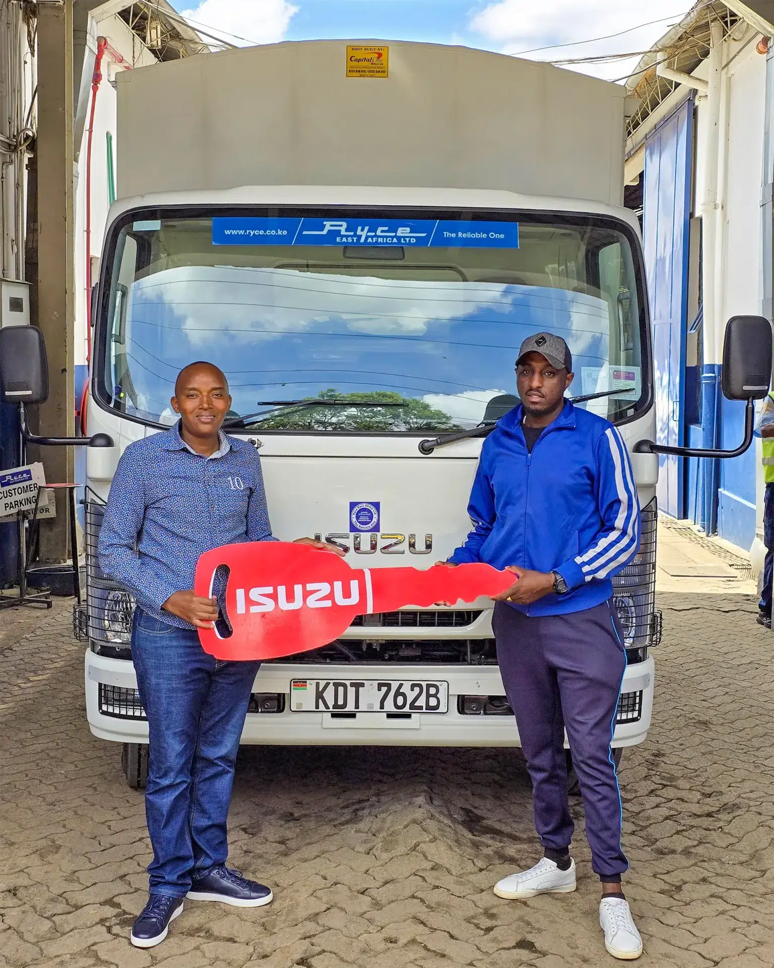 Wang'ombe, a top Isuzu sales representative, hands over a symbolic red Isuzu key to a representative of FARISI MANUFACTURING INDUSTRIES in front of a newly delivered Isuzu NQR 81 BLUEPOWER Truck at Ryce East Africa Ltd.