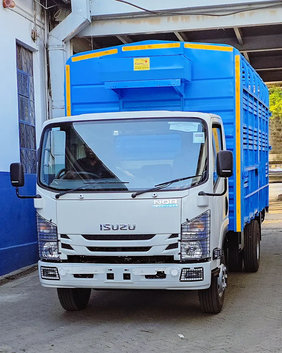 A front-three-quarter view of a white Isuzu NQR Blue Power truck equipped with a bright blue slatted livestock or multi-purpose cargo body. The body features yellow reflective safety tape along the edges and a "Capital" manufacturer label. The truck is shown with its hazard lights on, parked in an industrial loading area.