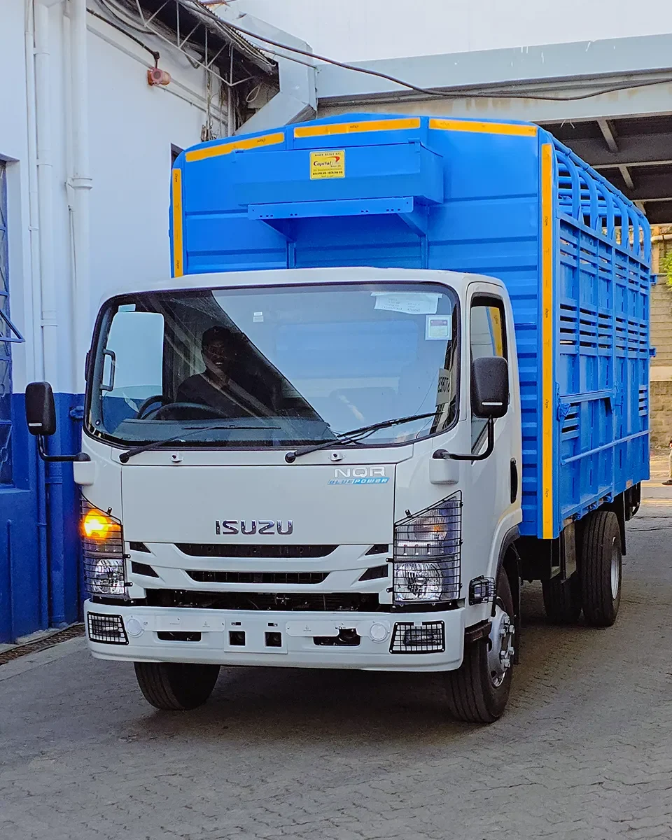 A front-three-quarter view of a white Isuzu NQR Blue Power truck equipped with a bright blue slatted livestock or multi-purpose cargo body. The body features yellow reflective safety tape along the edges and a "Capital" manufacturer label. The truck is shown with its hazard lights on, parked in an industrial loading area.
