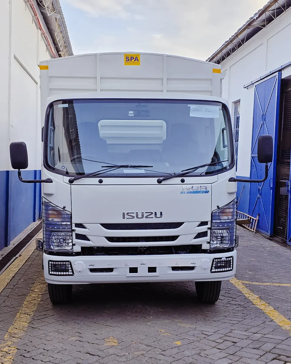 A front-angle view of a white Isuzu NQR Blue Power light-duty truck. It features a high-clearance cargo body built by SPA Industries Ltd, protective metal grilles over the headlights and fog lights, and a clean, industrial white finish. The truck is parked on a paved surface between two white buildings.