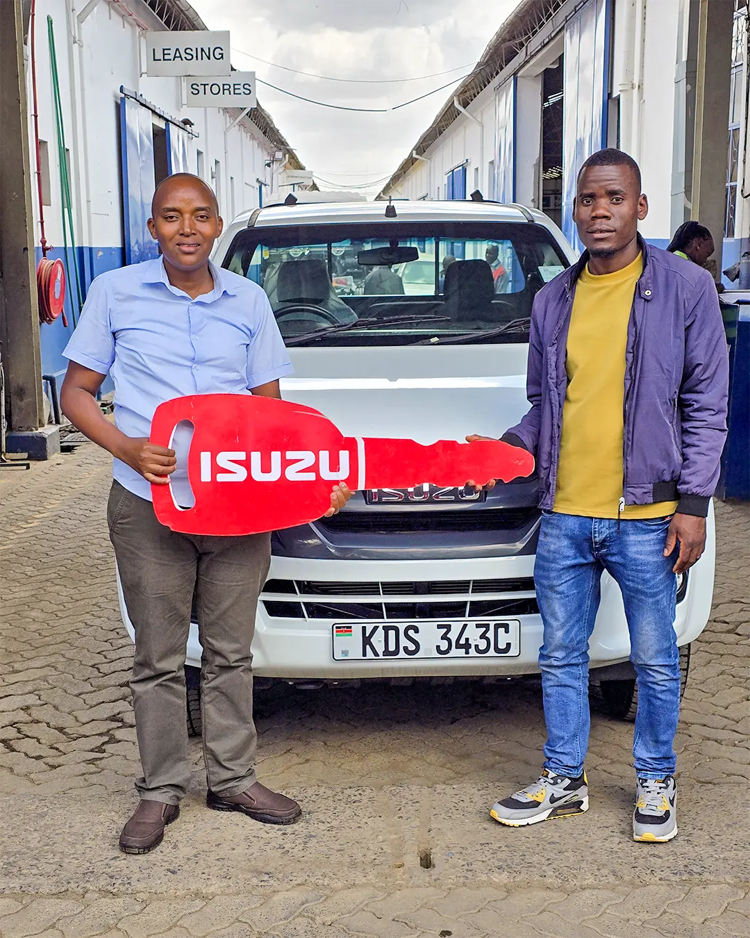 Two men standing in front of a white Isuzu D-Max Single Cab Pick-up holding a large red Isuzu key, marking the sale to Shiquenez Enterprises by Wang'ombe Truck Dealer.