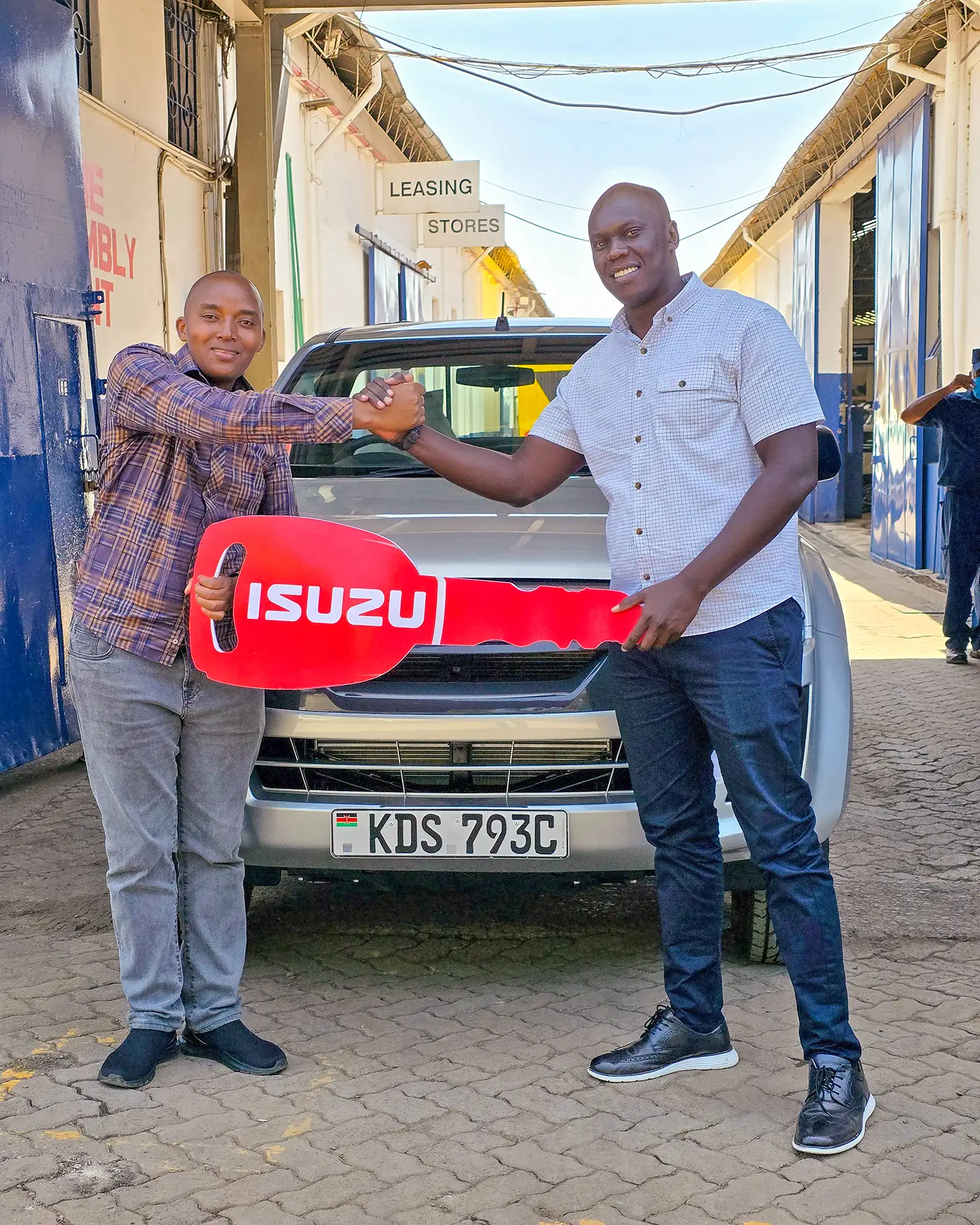 Wang'ombe, a top Isuzu sales representative, shaking hands with a satisfied customer from LIFESTYLE DRY CLEANERS while presenting the keys to a new Isuzu D-Max Single Cab truck. The truck is prominently displayed in the background with a visible license plate, "KDS 793C.