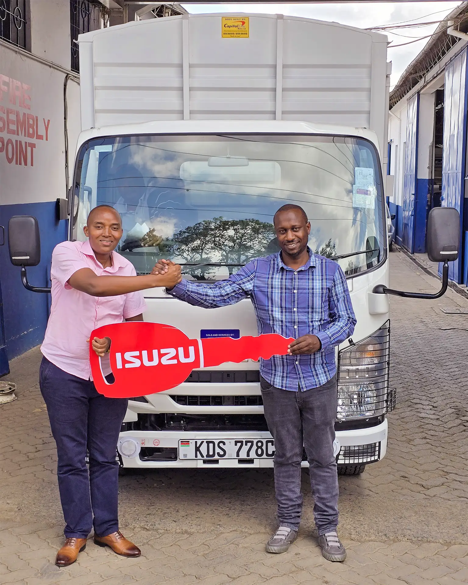 Wang'ombe, a top Isuzu sales representative in Kenya, handing over the keys to an Isuzu NMR 85 BLUEPOWER Truck to a satisfied Danshi Enterprises Ltd representative, in front of the newly purchased truck at a dealership in Nairobi.
